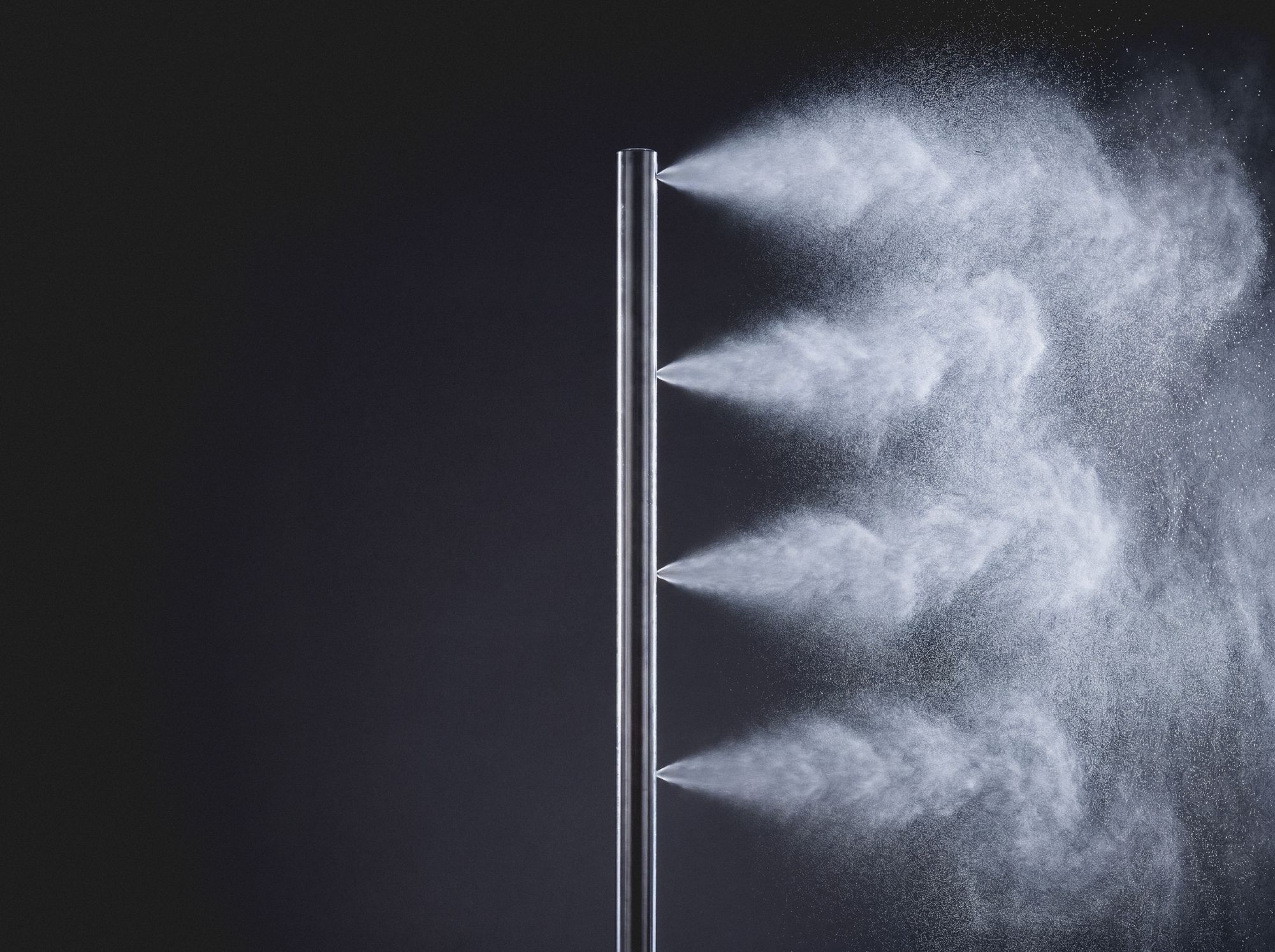 Studio detail of a Fontana misting pole in brushed stainless steel emitting four fine fog jets against a dark background